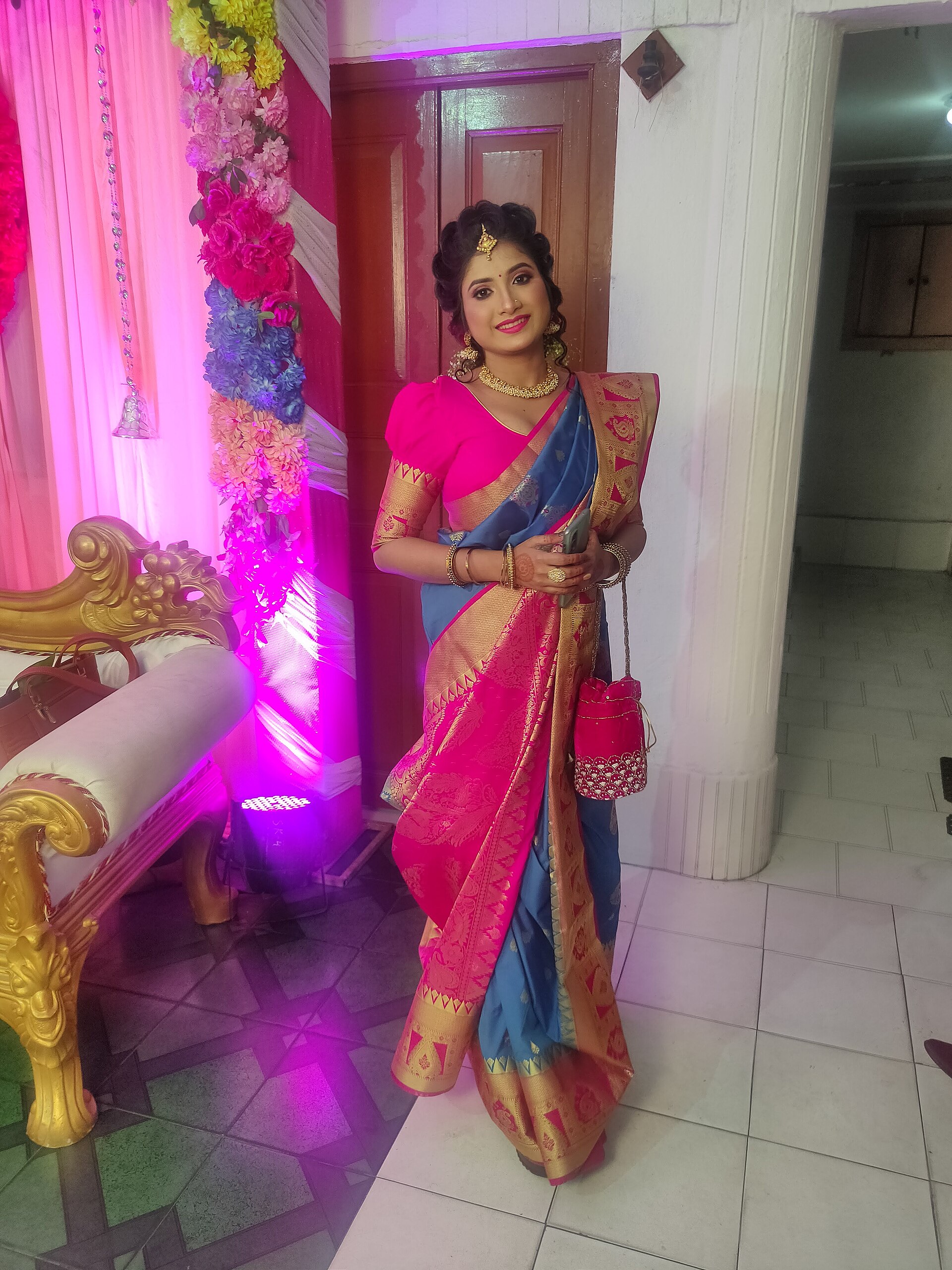 Saree | Stylo Aunty A woman showcases a vibrant pink and blue saree with gold jewelry, exemplifying desi fashion.